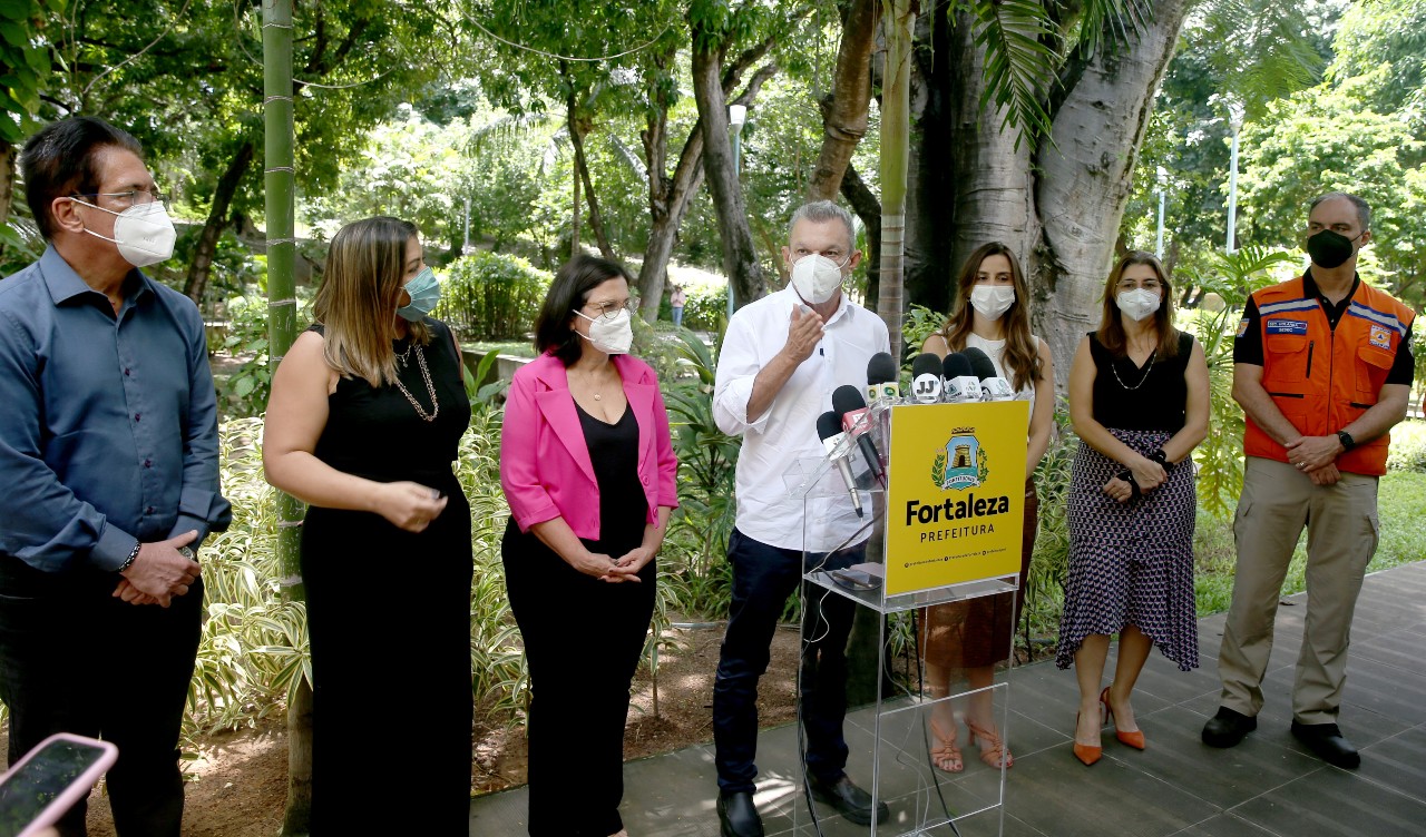 grupo de pessoas em pé em um jardim, todos usando máscara, enquanto dão entrevista